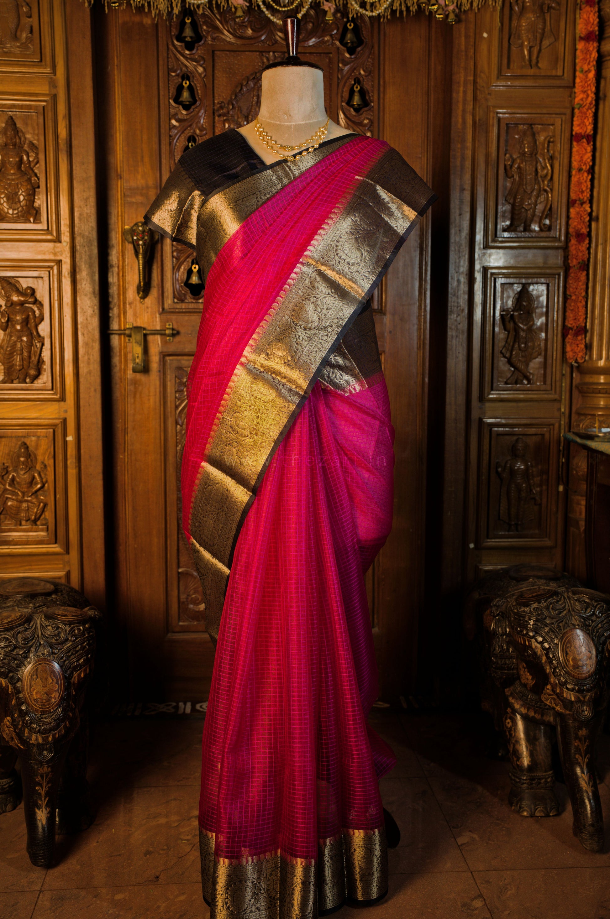Mannequin wearing a pink saree with gold and silver border against a decorative wooden background